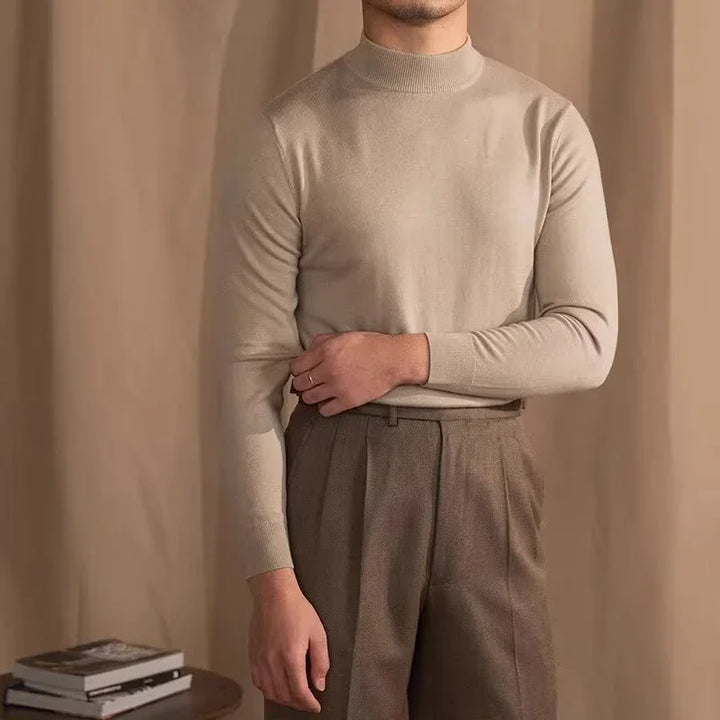 Man wearing beige turtleneck sweater and brown pleated trousers against brown backdrop