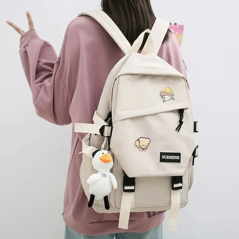 Beige backpack with cartoon pins and plush duck keychain worn by person in pink sweatshirt