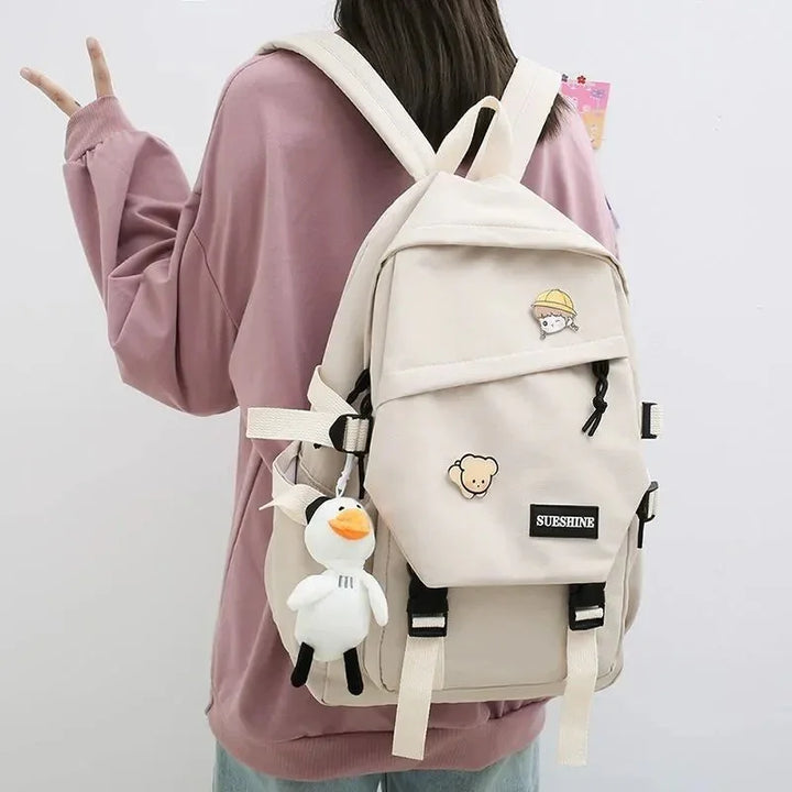 Beige backpack with cartoon pins and plush duck keychain worn by person in pink sweatshirt
