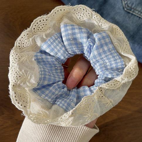 Hand holding a blue and white gingham scrunchie with lace trim over a wooden surface