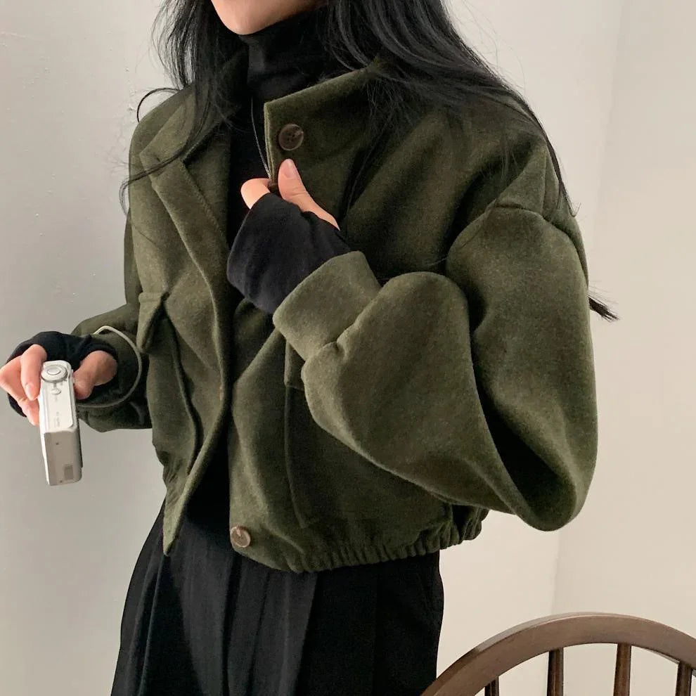 Woman wearing a cropped olive green jacket over black turtleneck and holding a white camera indoors