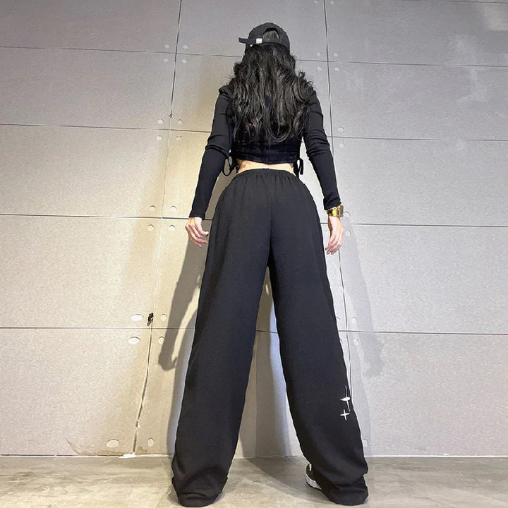 Woman in black wide-leg pants, black long sleeve crop top, and cap standing against tiled wall