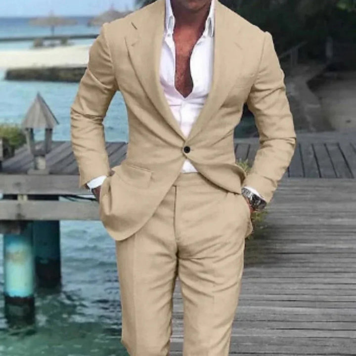 Man wearing beige suit and white shirt, standing on wooden pier by water with hands in pockets