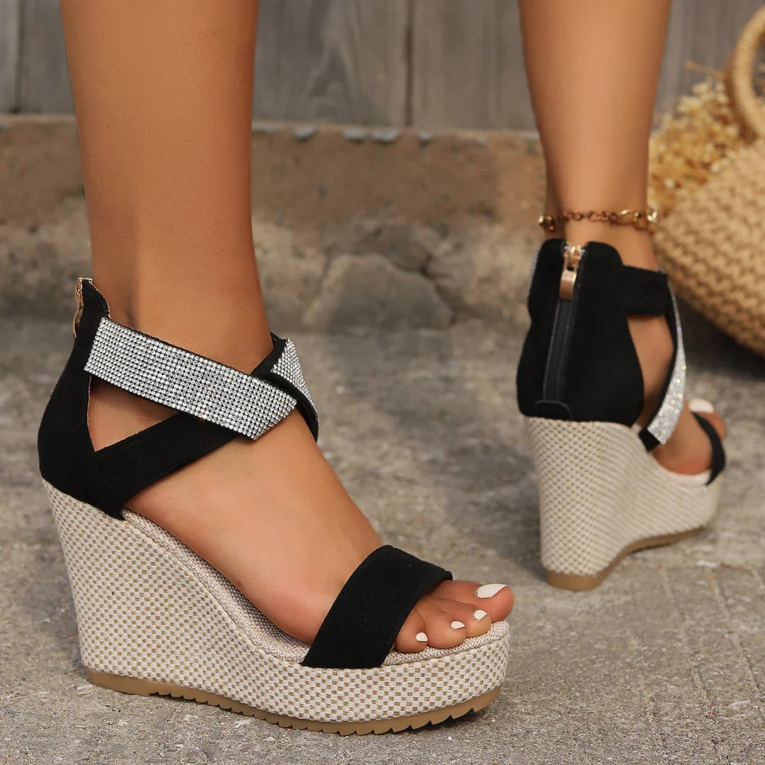 Black wedge sandals with rhinestone ankle straps on textured concrete floor