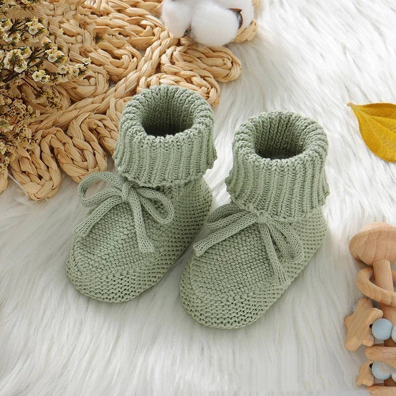 Soft green knitted baby booties with ties on white furry surface, surrounded by dried flowers and wooden toys