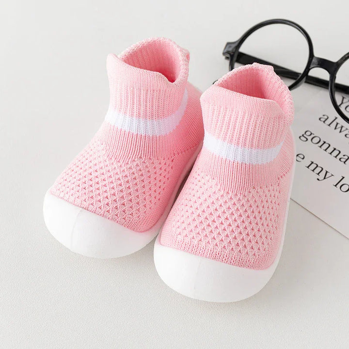 Pink knitted baby shoes with white toe caps and a white stripe, placed next to black glasses