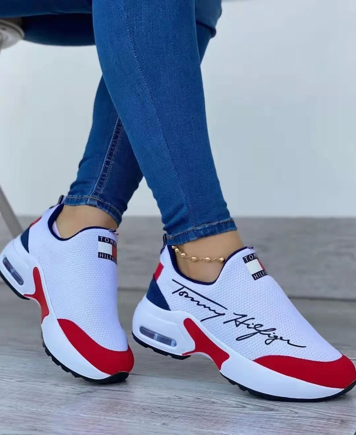 Person wearing white, red, and navy Tommy Hilfiger sneakers with blue jeans on wooden floor
