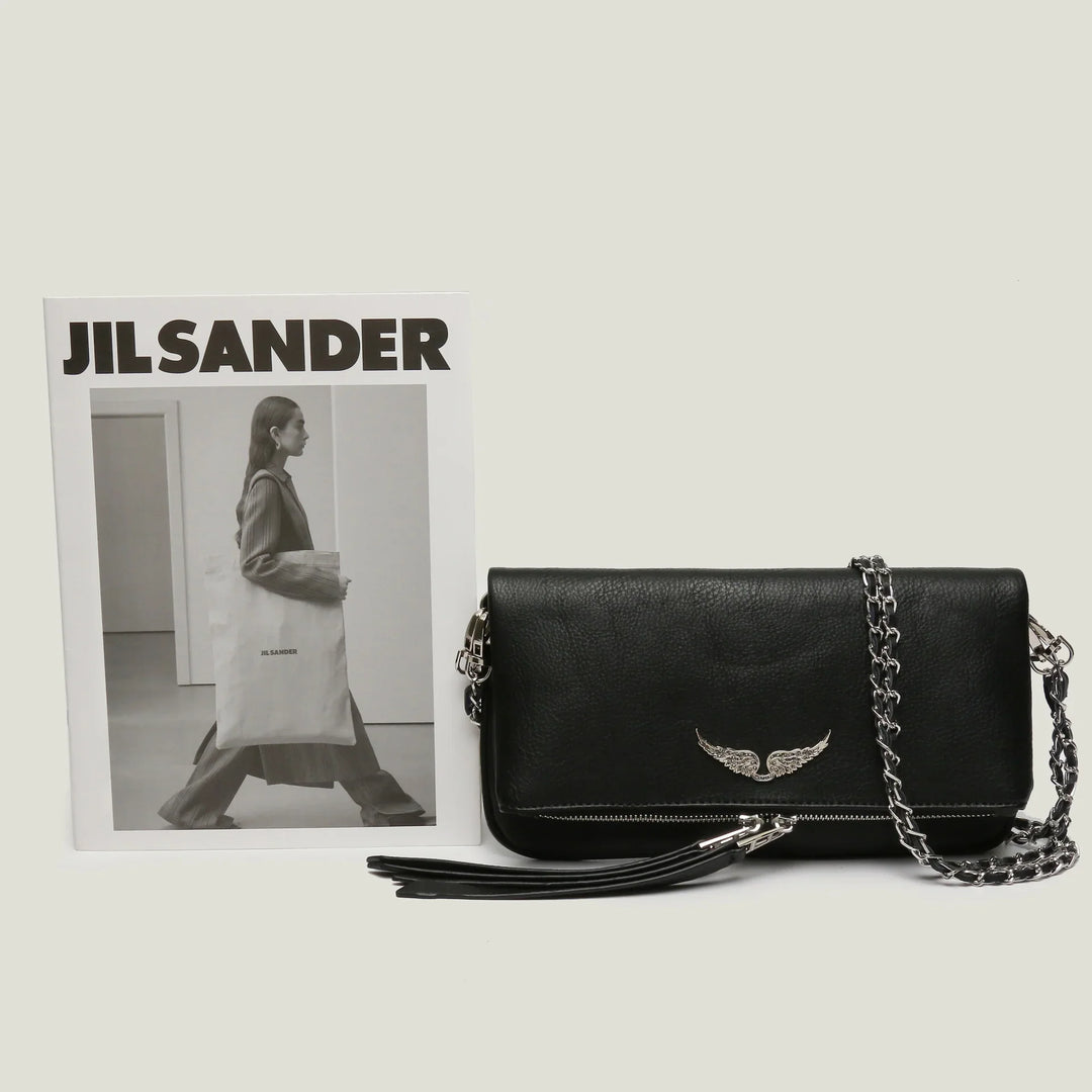 Black leather shoulder bag with silver wing emblem and chain strap beside Jil Sander black and white fashion magazine