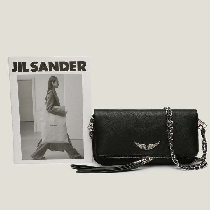 Black leather shoulder bag with silver wing emblem and chain strap beside Jil Sander black and white fashion magazine