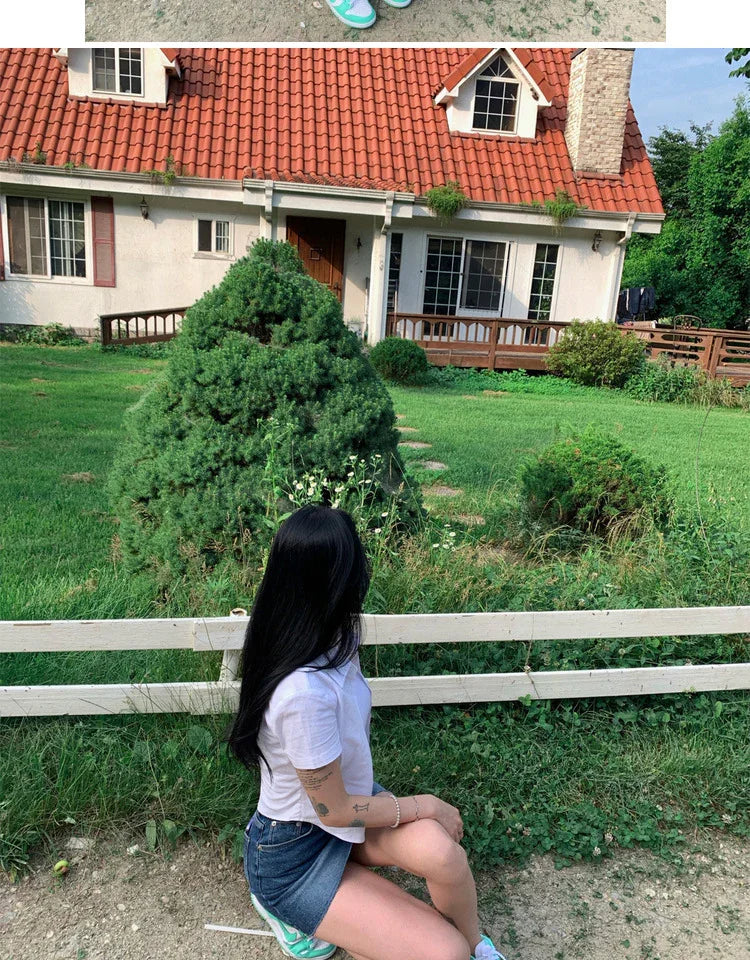 Woman with long black hair in white shirt and denim shorts sitting near green bushes outside a house with red roof