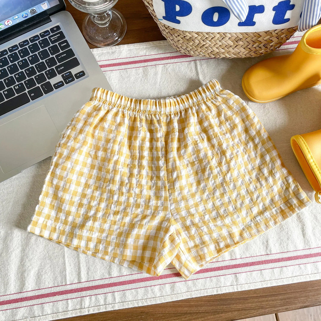 Yellow and white checkered cotton shorts on a table with laptop, glass, wicker bag, and yellow rain boots