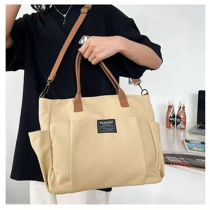Beige fashion tote bag with brown handles and adjustable shoulder strap held by person in black shirt