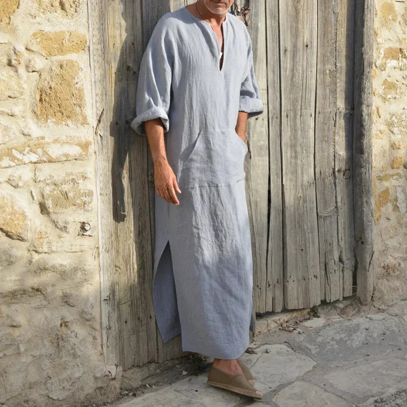 Man wearing light gray linen long tunic with rolled sleeves and side slits standing against rustic wooden door