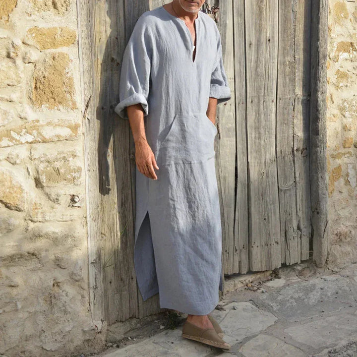 Man wearing light gray linen long tunic with rolled sleeves and side slits standing against rustic wooden door