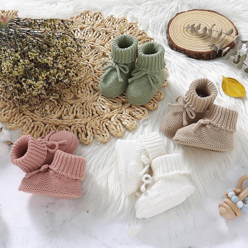 Four pairs of knitted baby booties in green, beige, pink, and white displayed on a soft white surface with rustic decor
