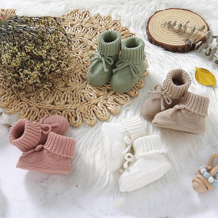 Four pairs of knitted baby booties in green, beige, pink, and white displayed on a soft white surface with rustic decor