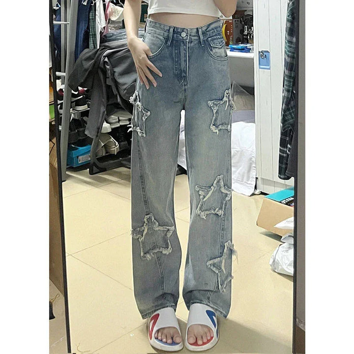 Person wearing high-waisted denim jeans with frayed star patches and white sliders in a bedroom