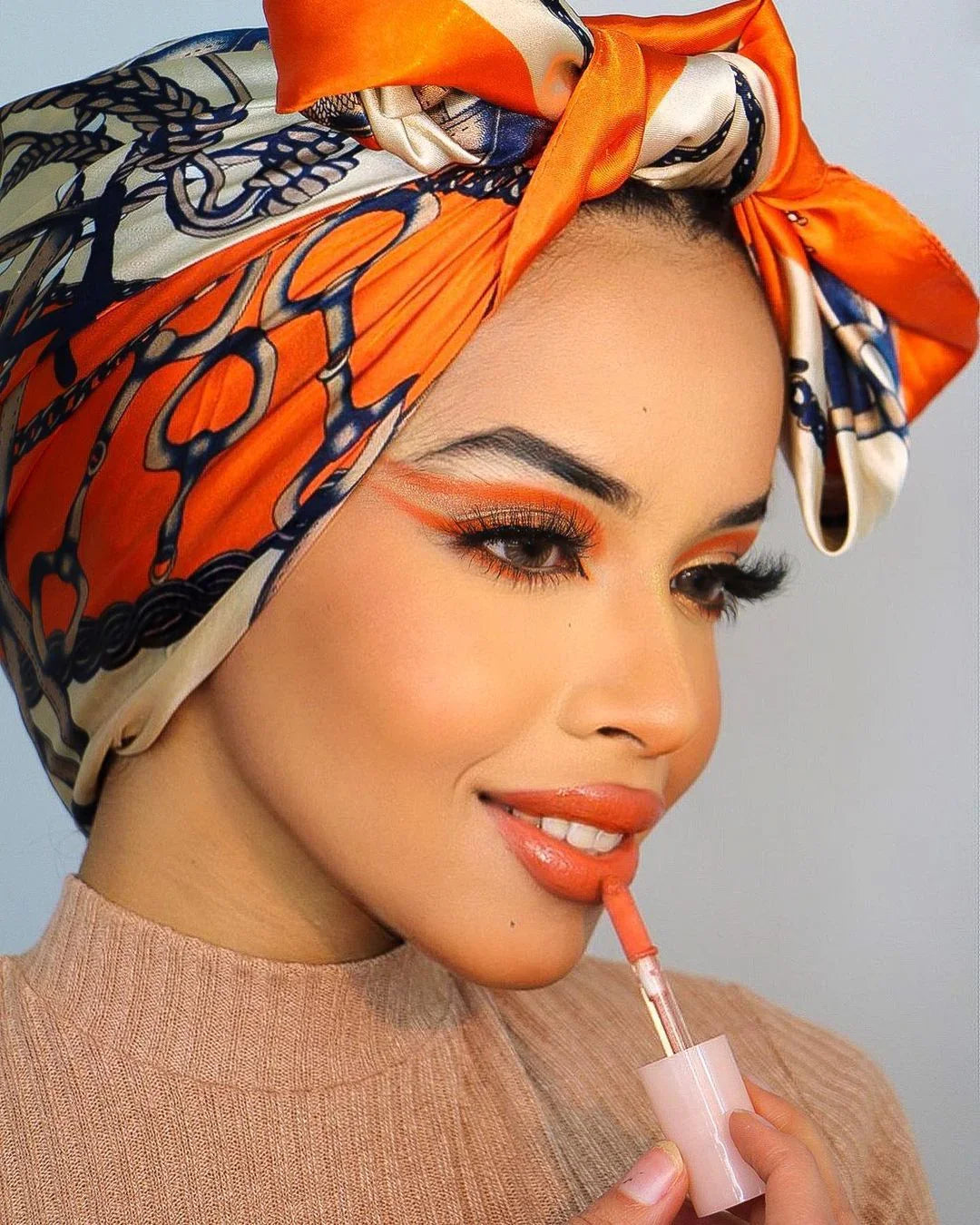 Woman wearing an orange and blue patterned headscarf applying coral lipstick with matching eye makeup