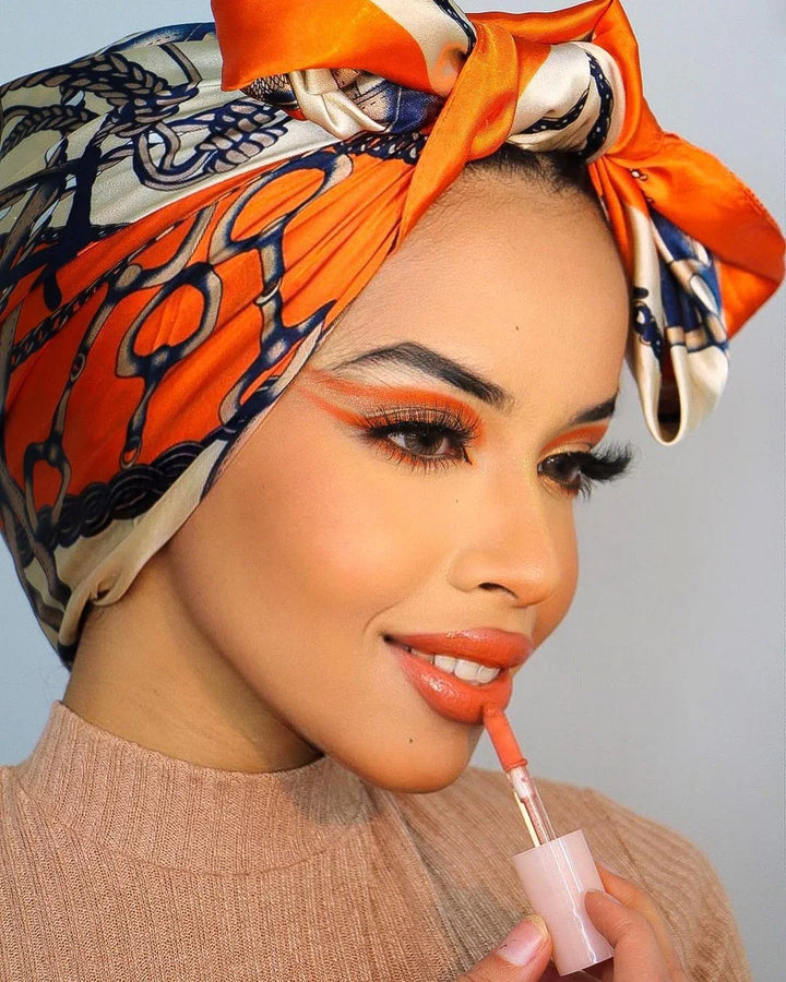 Woman wearing an orange and blue patterned headscarf applying coral lipstick with matching eye makeup