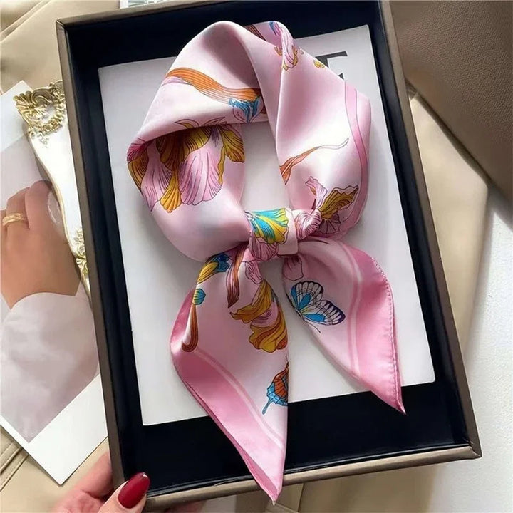 Pink silk scarf with floral and butterfly print displayed in black gift box on a beige surface