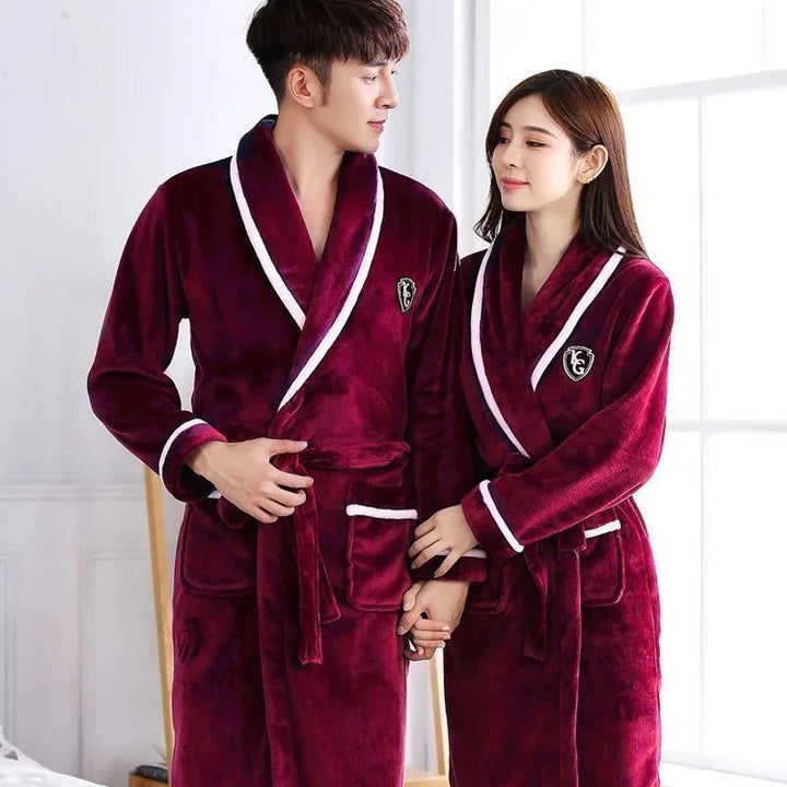 Couple wearing matching burgundy plush bathrobes with white trim, holding hands indoors by window