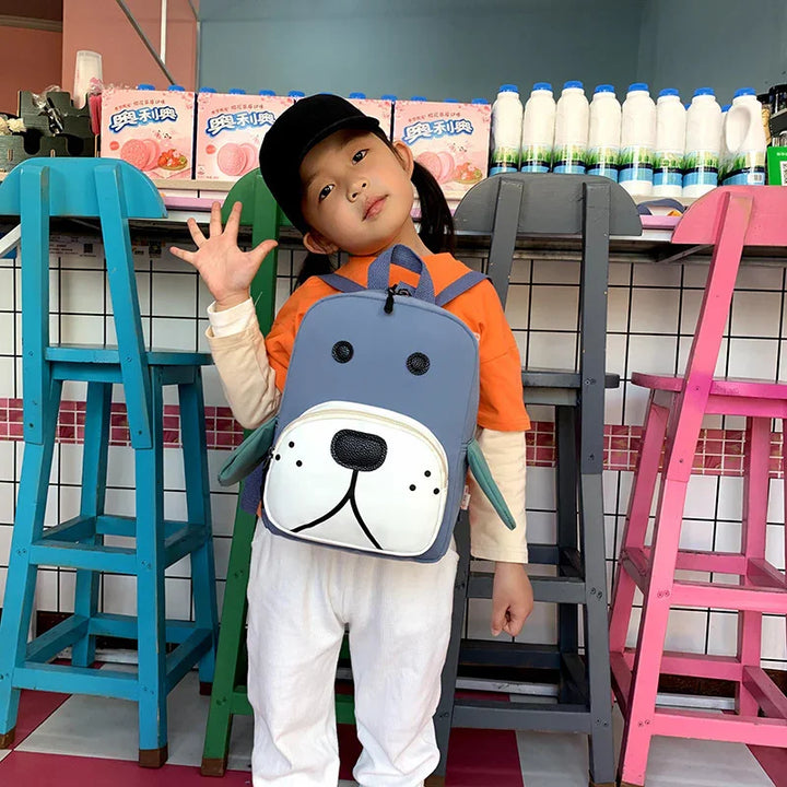 Child wearing black cap and orange shirt with gray dog face backpack standing by colorful chairs in cafe