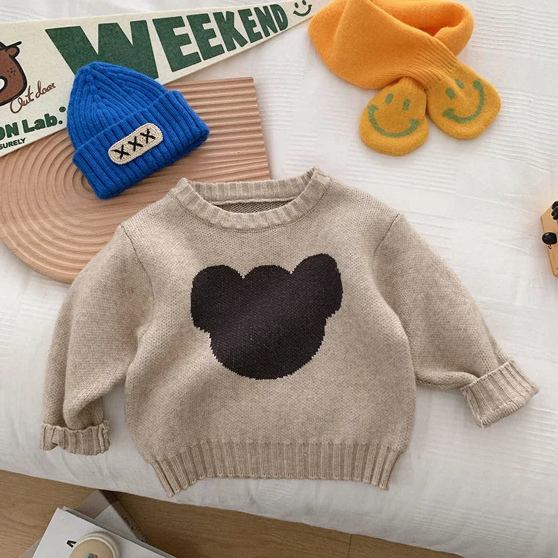 Beige kids sweater with black bear silhouette, blue knit hat, orange socks with smiley faces on white bed