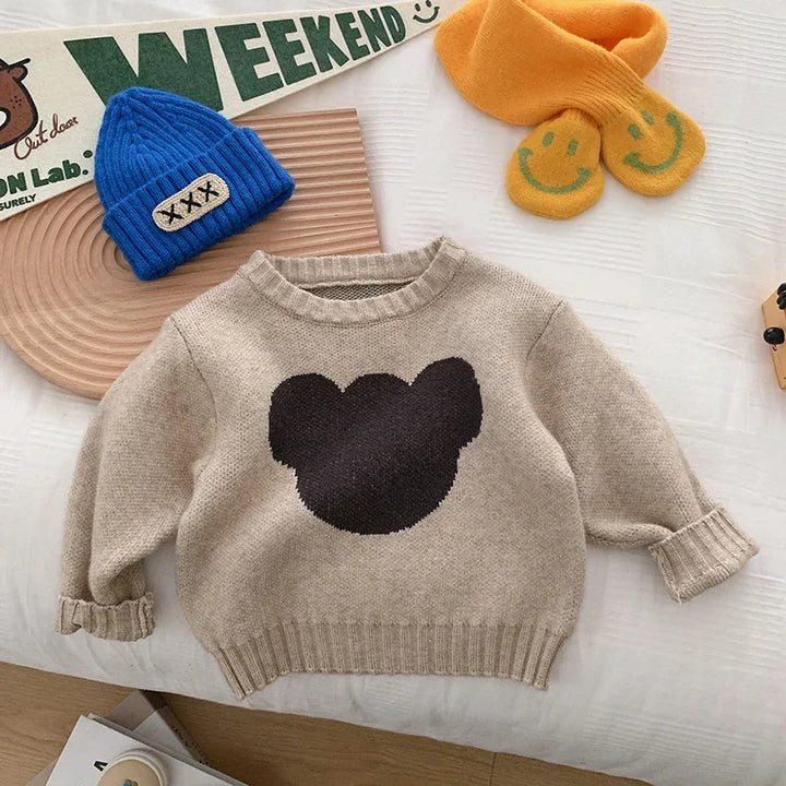 Beige kids sweater with black bear silhouette, blue knit hat, orange socks with smiley faces on white bed