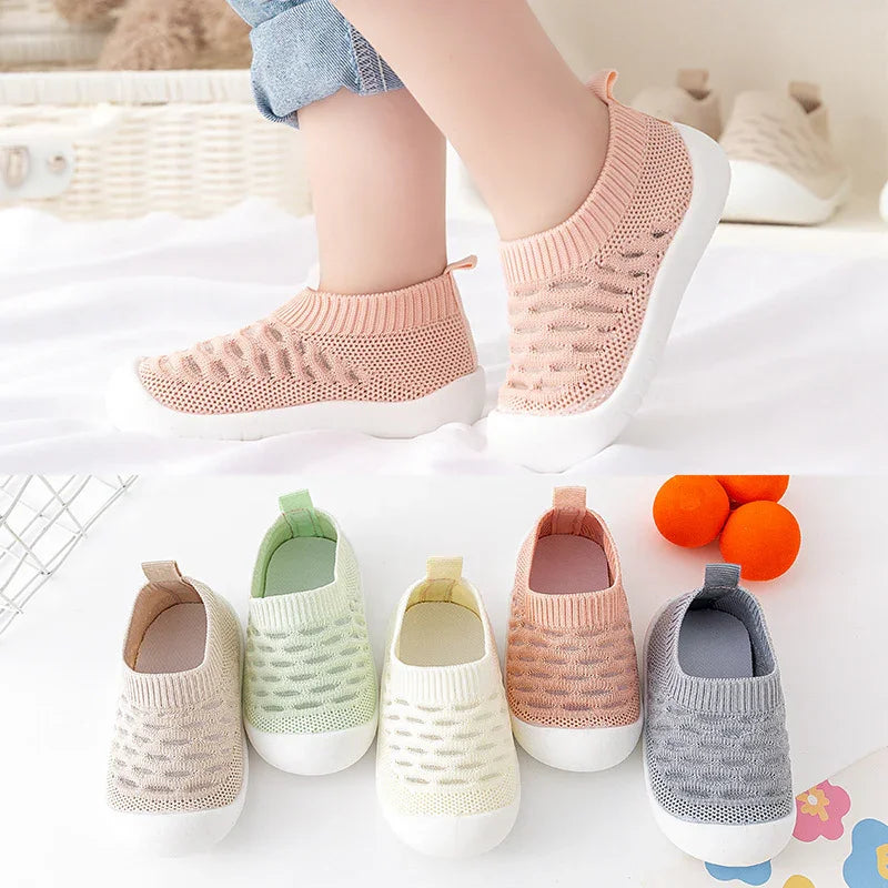 Child wearing pink knitted slip-on shoes with white soles, surrounded by similar shoes in pastel colors on a white surface