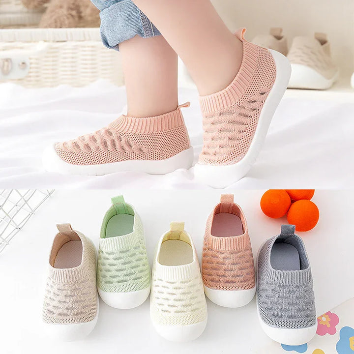 Child wearing pink knitted slip-on shoes with white soles, surrounded by similar shoes in pastel colors on a white surface