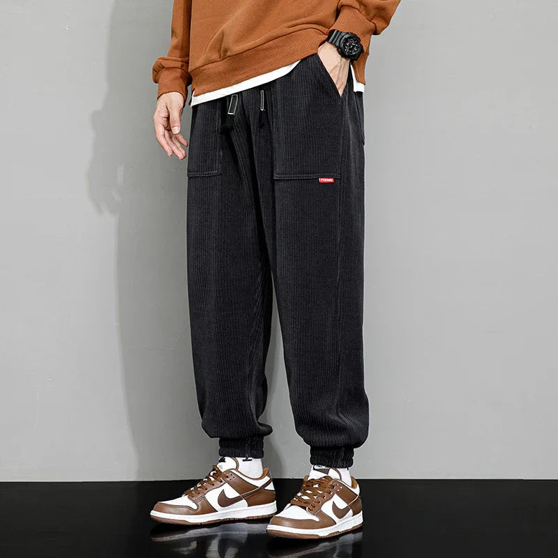 Man wearing black corduroy jogger pants with patch pockets and brown-white Nike sneakers on a gray background