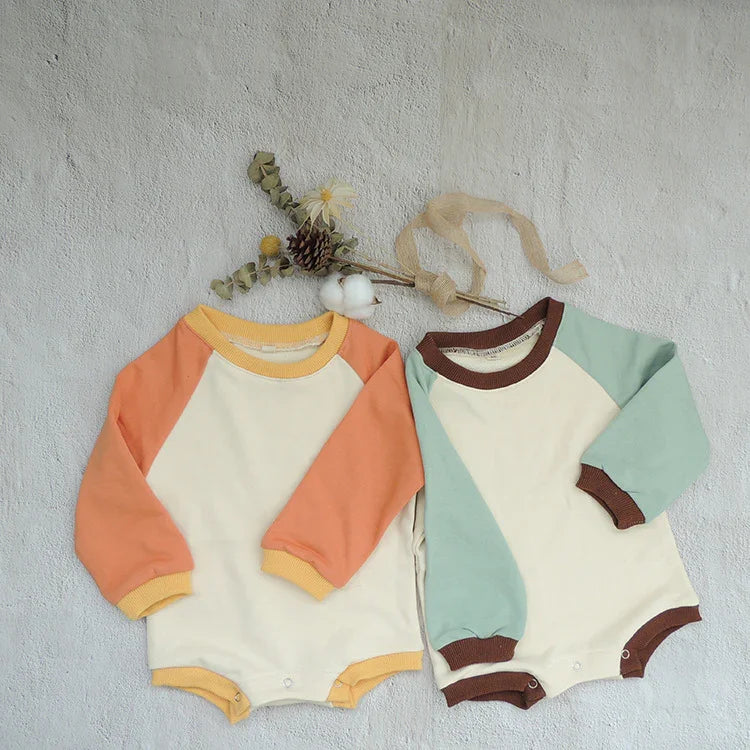 Two long-sleeve baby rompers with raglan sleeves in pastel orange and mint green, laid flat on textured surface with dried flowers