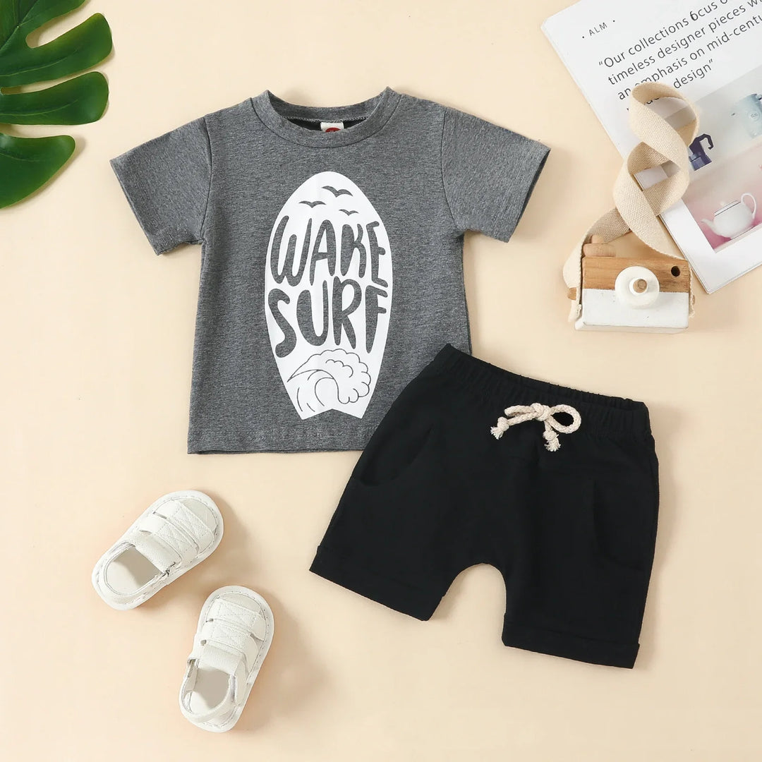 Toddler outfit with grey 'Wake Surf' graphic t-shirt, black shorts with drawstring, and white baby shoes on beige background