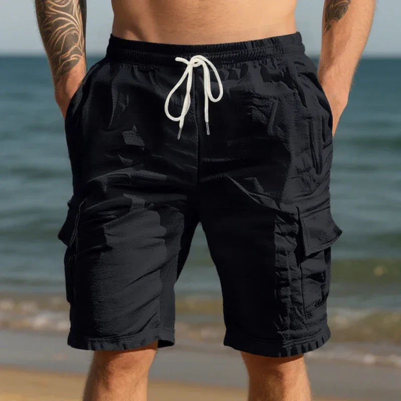 Man wearing black cargo shorts with white drawstring standing on beach with ocean background