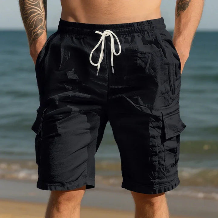 Man wearing black cargo shorts with white drawstring standing on beach with ocean background