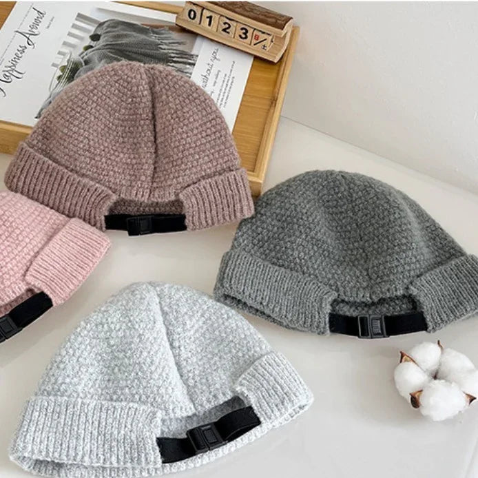 Three knitted beanie hats in pink, gray, and light gray on a white surface with a wooden tray and cotton flower