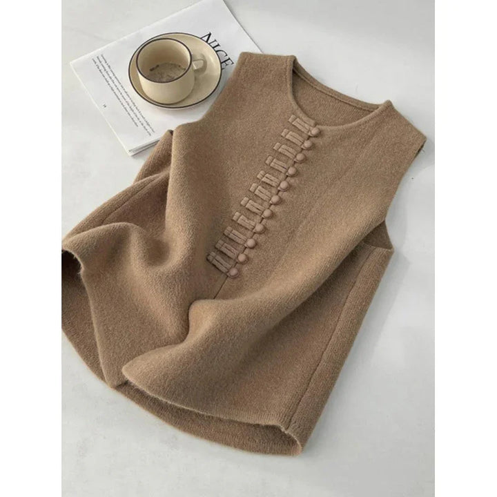 Beige sleeveless wool vest with decorative buttons laid on white surface next to beige cup and magazine