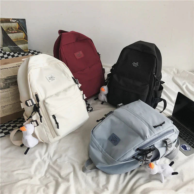 Four backpacks in white, red, black, and gray with duck keychains on a bed next to a laptop