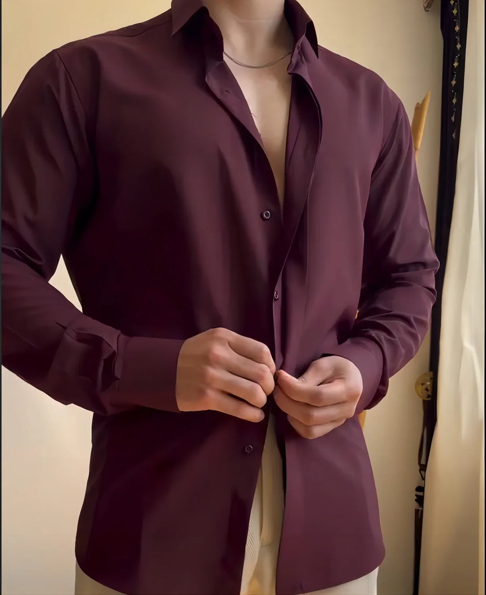 Man buttoning a dark maroon dress shirt with beige pants indoors by a window curtain