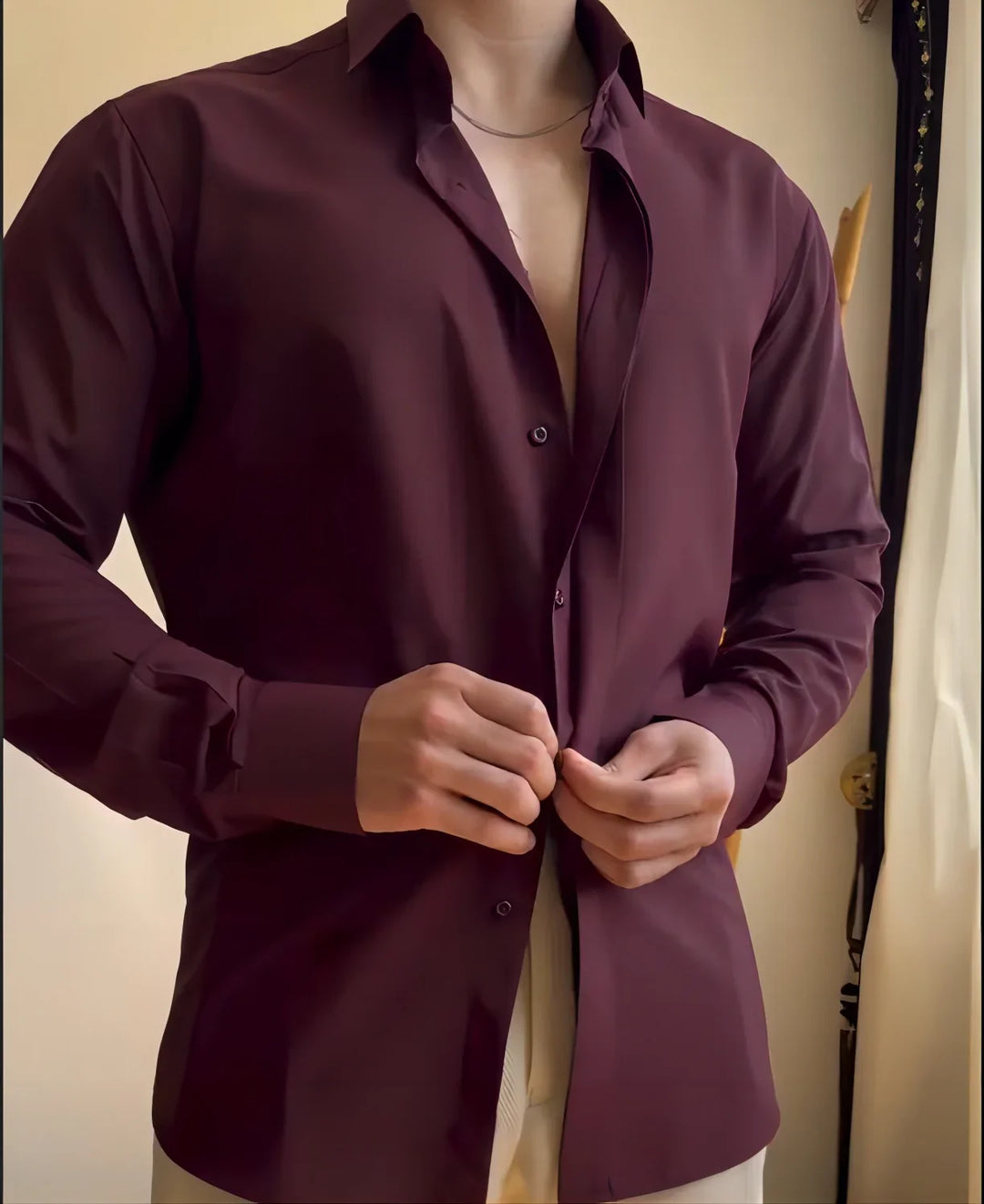 Man buttoning a dark maroon dress shirt with beige pants indoors by a window curtain