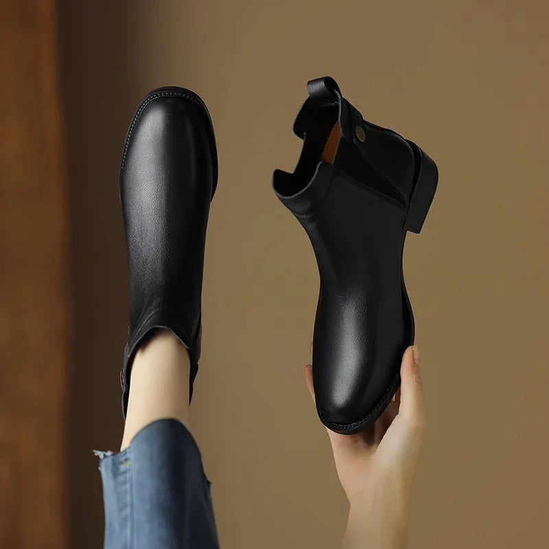 Black leather ankle boots with pull tabs, one on foot and one held by hand against neutral background
