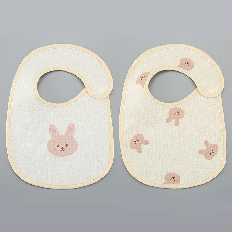 Two cream baby bibs with beige trim; one features a single pink bunny face, the other multiple pink bunny faces