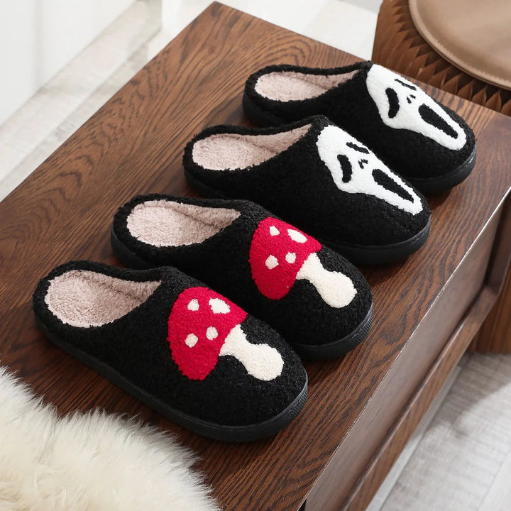 Two pairs of black fuzzy slippers on wooden surface, one pair with red mushroom design, one with white ghost face