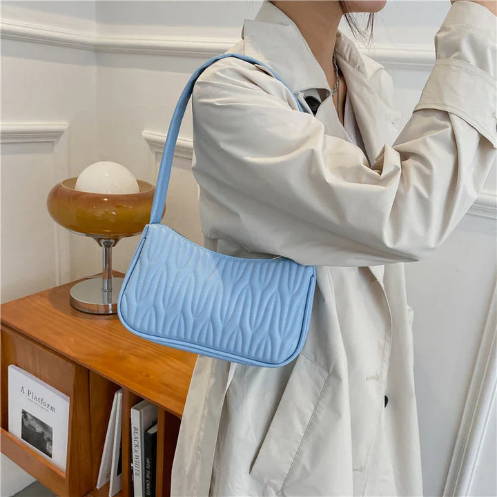 Woman wearing beige trench coat with light blue quilted shoulder bag in modern indoor setting