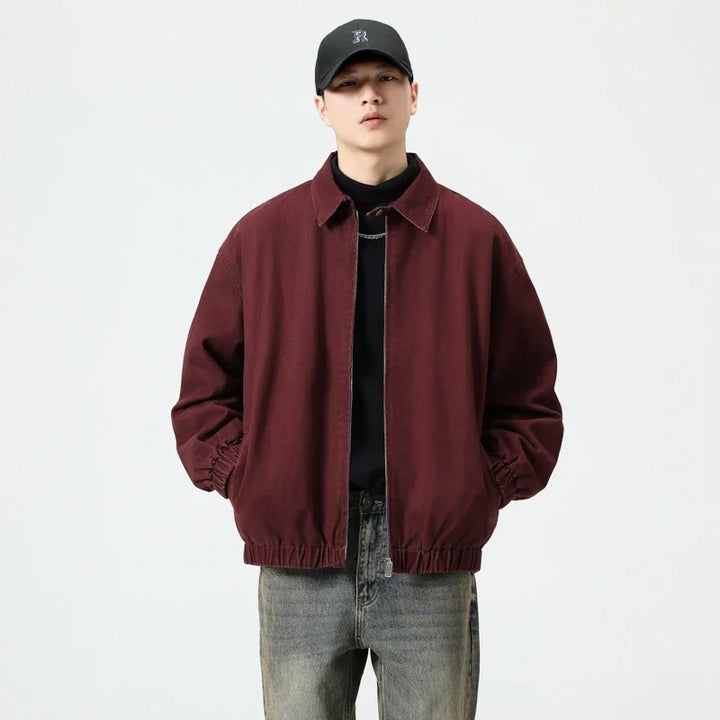 Man wearing maroon casual jacket with elastic cuffs, gray jeans, black shirt, and black cap