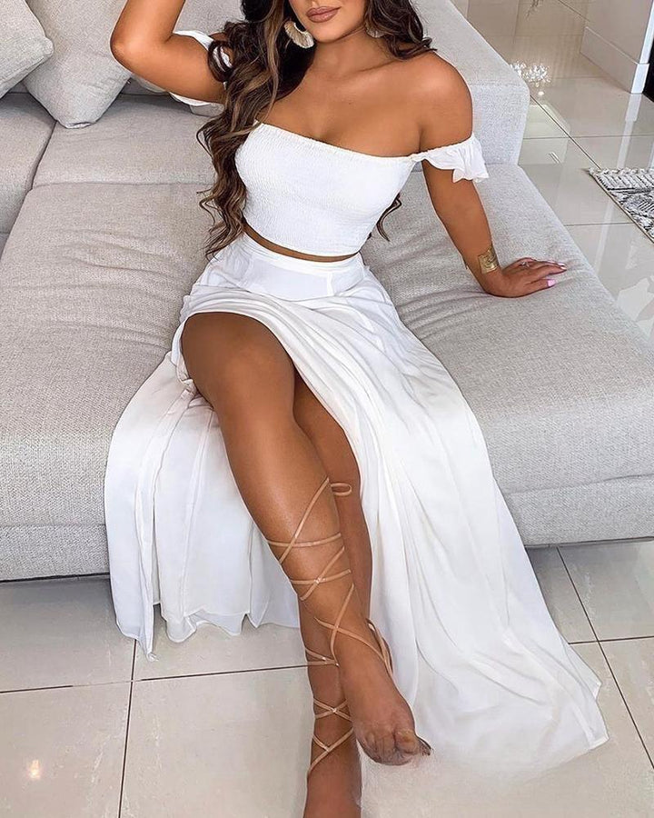 Woman sitting on light gray sofa wearing white off-shoulder crop top and flowing white skirt with lace-up sandals