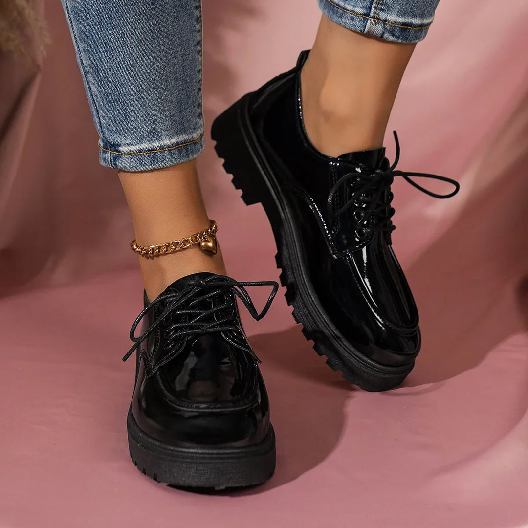 Black glossy platform shoes with thick soles worn with rolled-up jeans and gold heart anklet on pink background