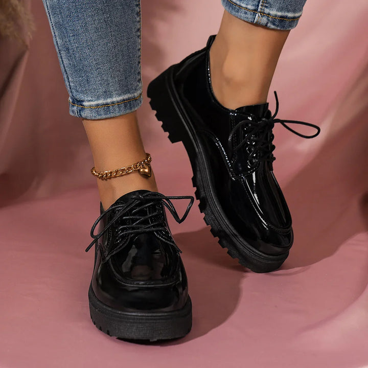 Black glossy platform shoes with thick soles worn with rolled-up jeans and gold heart anklet on pink background