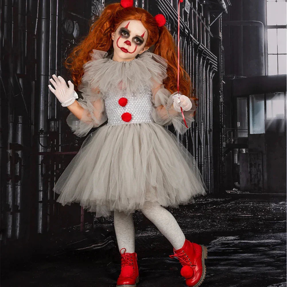 Child in gray tulle clown costume with red pom poms and red boots holding a red balloon in dark industrial setting