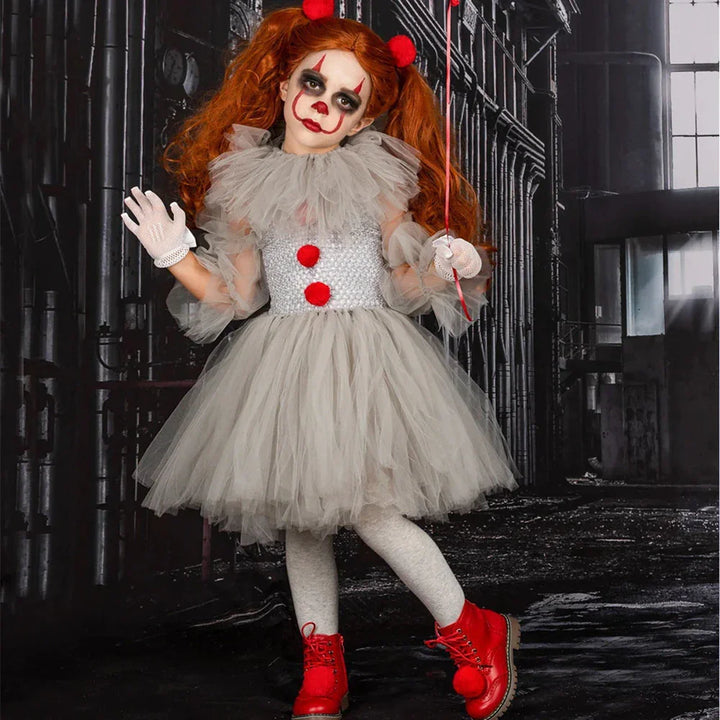 Child in gray tulle clown costume with red pom poms and red boots holding a red balloon in dark industrial setting